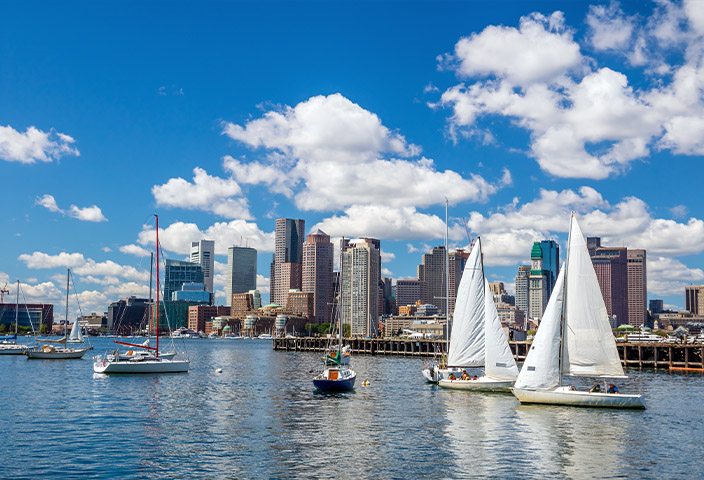 Boston Harbor skyline view with sailboats on blue water under a cloudy sky.
