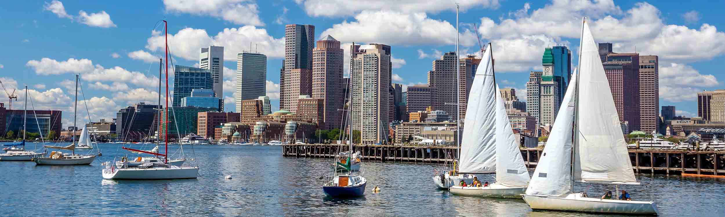 Boston Harbor skyline view with sailboats on blue water under a cloudy sky.